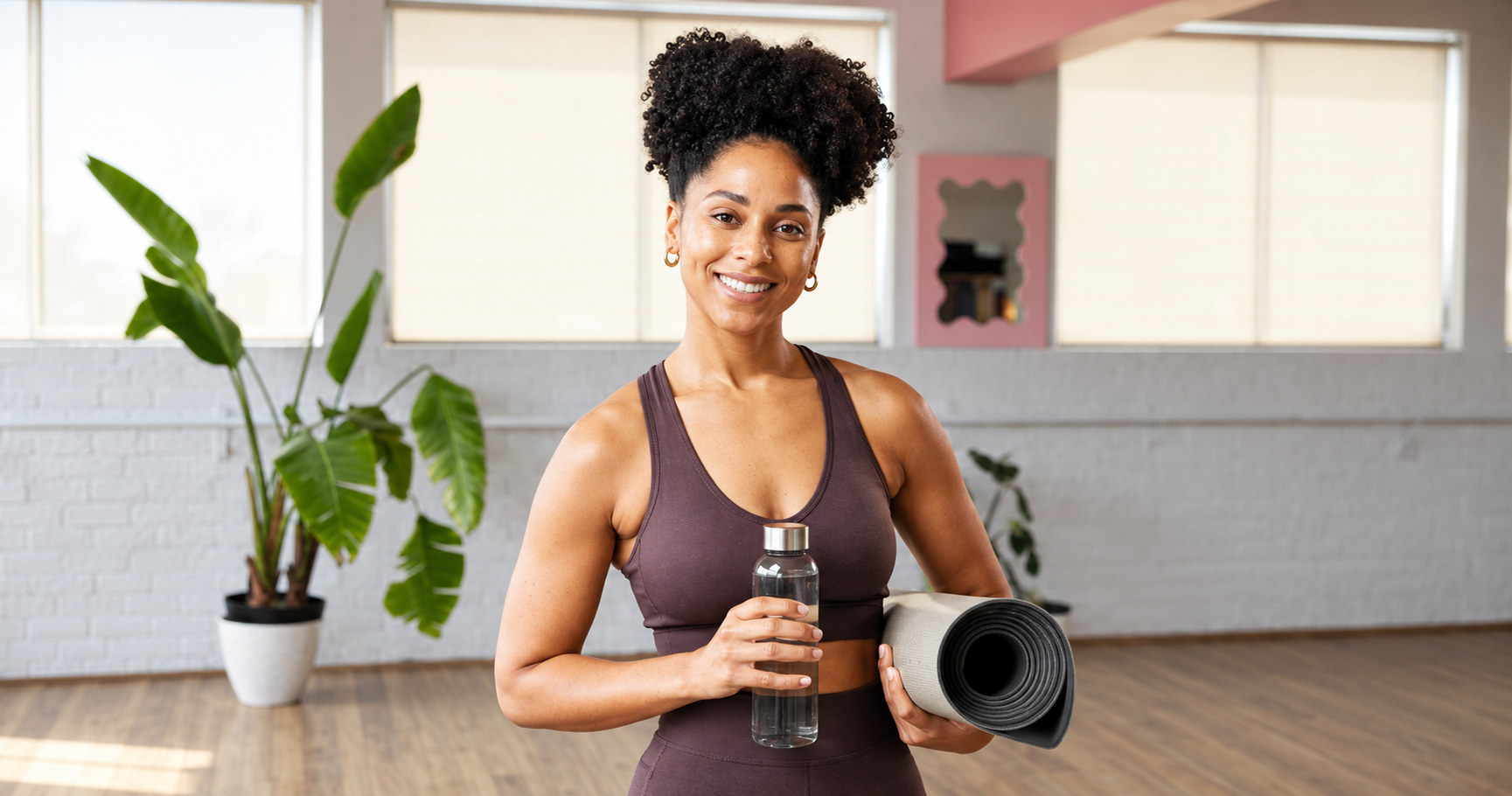 A woman smiling in a yoga studio, holding a mat and a water bottle